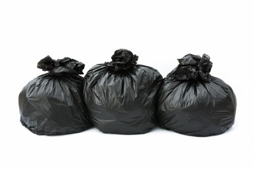 Three black rubbish bags made of plastic, tightly knotted at the top, are aligned in a row against a plain white background. The bags are full and appear to contain garden waste or general rubbish, with slight creases and a matte surface texture. The middle bag is positioned slightly in front of the others, with the left and right bags leaning slightly inward. The clear, minimalistic background accentuates the bags, emphasizing their size and shape. This image visually represents waste collection or rubbish removal services, such as those provided by Garden Clearance Seven Sisters, which may operate within the London borough or surrounding area. The depiction highlights the standard form and appearance of waste bags used for garden and household clearance tasks in the local context.