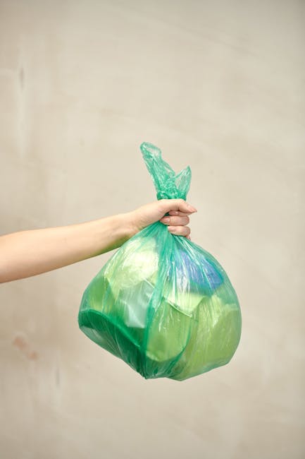 A person's arm is extended into the frame, holding a green plastic bag tied at the top with visible twists of the bag's opening. The bag appears full and slightly bulging, with a smooth, shiny surface