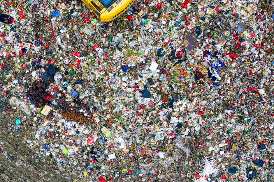 A crumpled red Coca-Cola can lies partially buried on a sandy surface, with its top dented and the label faded, surrounded by fine, textured light brown sand. The can's surface is shiny with visible s