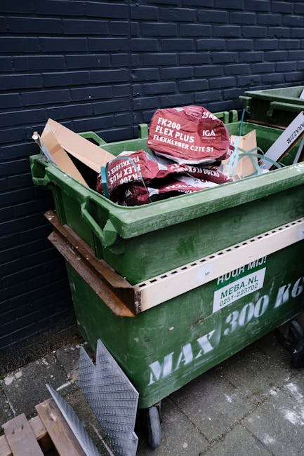 A waste collection worker, dressed in a high-visibility orange and yellow uniform, is standing behind a large red rubbish truck with a hydraulic waste compactor at the rear, positioned on the side of 
